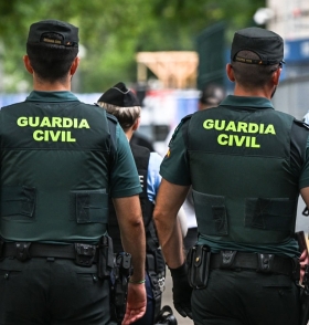 Two Guardia Civil officers, seen from behind, wear dark green uniforms with "GUARDIA CIVIL" in yellow on their backs.