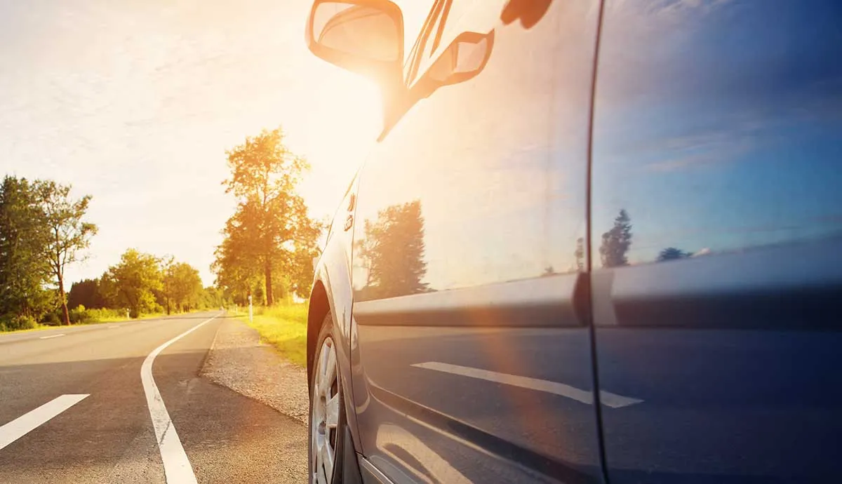 Close-up of a car's side with sunlight flaring over the side mirror and blue sky reflected in the window.