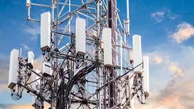 Close-up of a telecommunications tower, featuring multiple white rectangular antennas and a complex network of black cables, set against a cloudy sky.