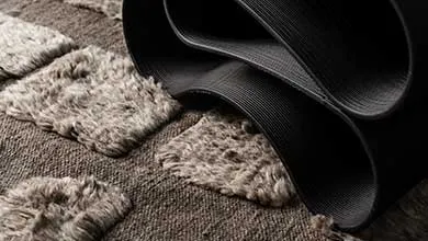 Close-up of a brown, textured rug with raised, shaggy gray squares. One side is rolled, revealing a black, ribbed, non-slip backing.