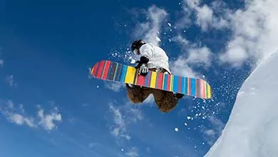 Snowboarder in mid-air against a blue sky, helmeted, with a colorful, striped board.