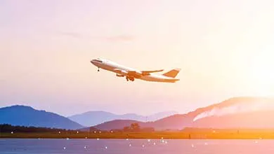 A passenger airplane takes off from a runway at sunrise or sunset, with mountains in the background and the sky glowing with warm light.
