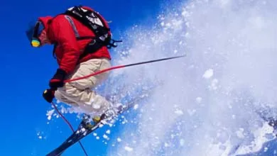 A skier launching off a snowy slope, creating a spray of snow against a clear blue sky.