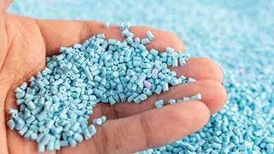 Hand holding small light-blue plastic pellets, with a larger pile of pellets in the background.