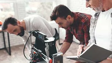 Two men examine a 3D printer while a third man stands next to them, writing in a notebook.
