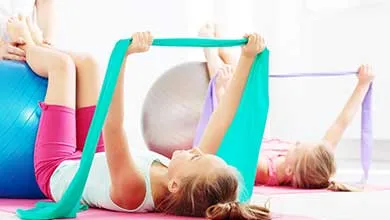 Two young girls lie on pink yoga mats, stretching resistance bands over large exercise balls.