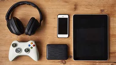 Overhead view of black headphones, a white game controller, a smartphone, and a tablet arranged on a wooden surface.